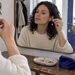 Woman styling two mismatched earrings at mirror